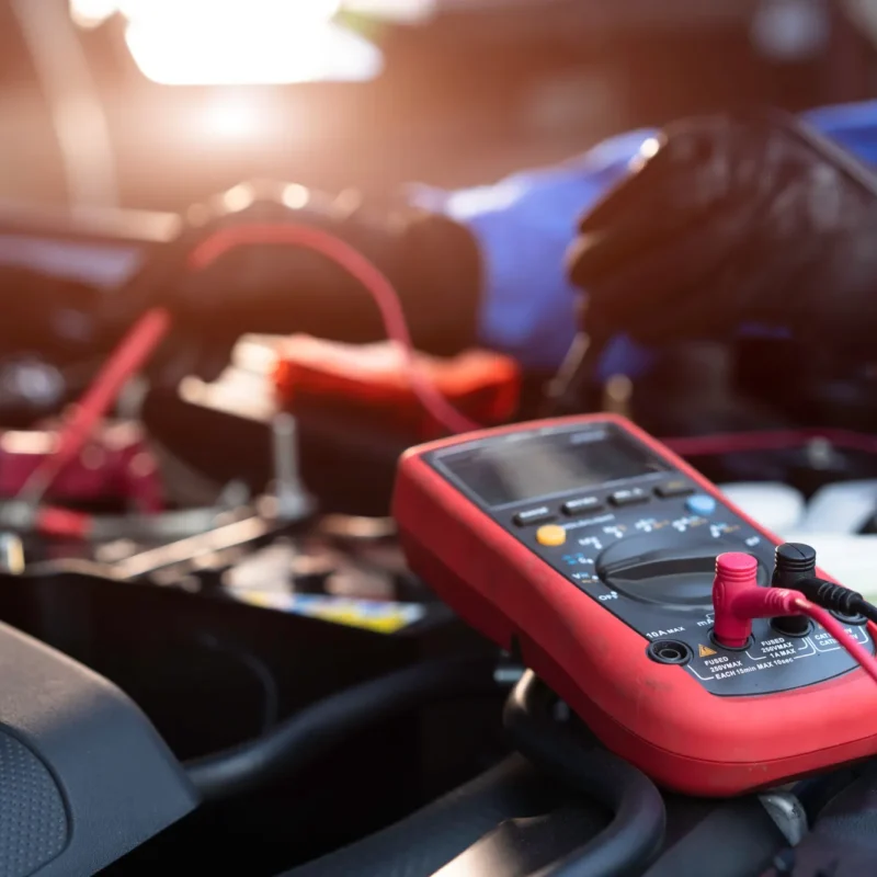 A red digital multimeter with red and black test leads sits on a car engine while a gloved technician works in the background.