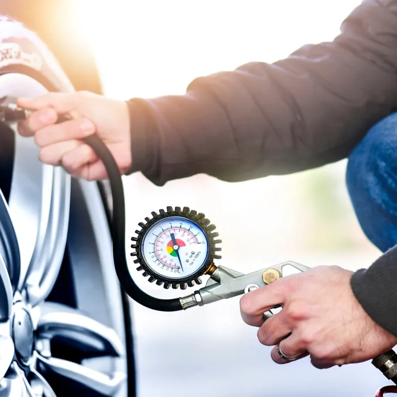 A man kneeling next to a car wheel checks the tire's air pressure with a hose-connected pressure gauge.