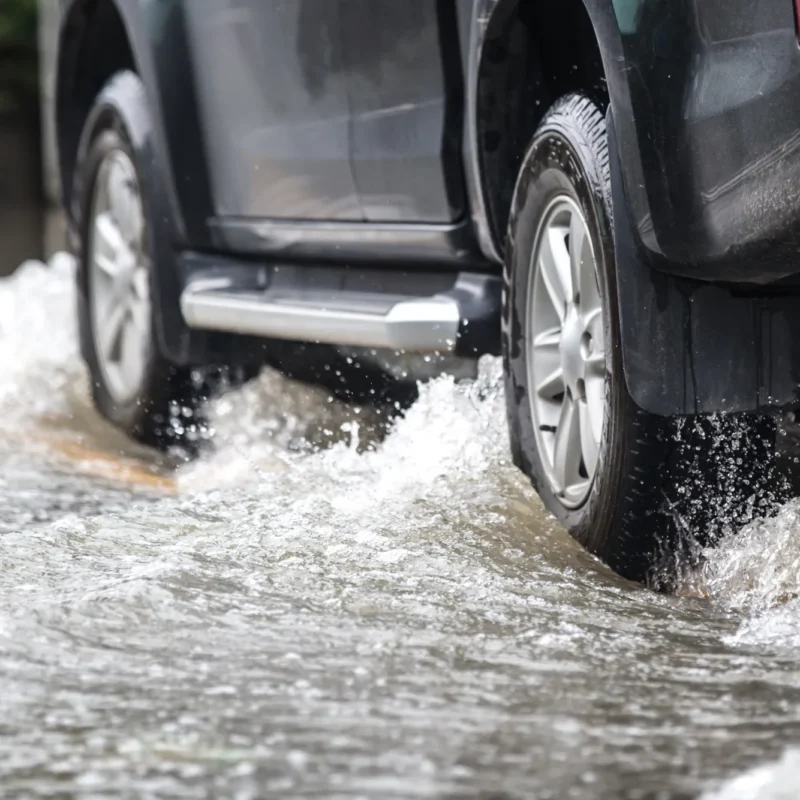 Rear tire and wheel of a dark SUV splashing through deep water on a flooded road.