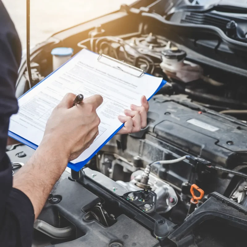 A mechanic holding a clipboard with a service inspection form while standing over a car's open engine bay.