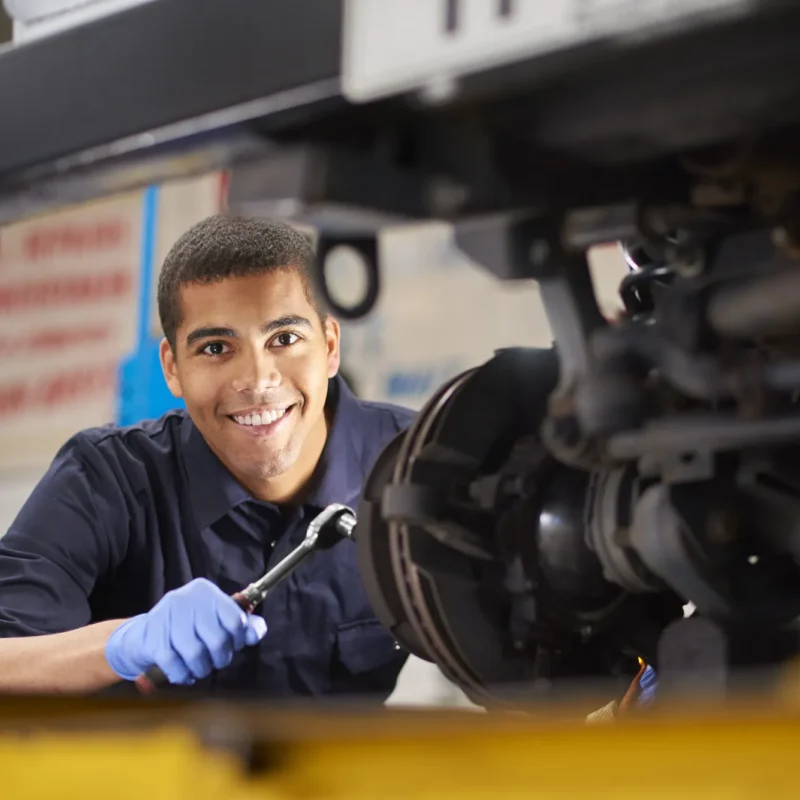 A smiling mechanic in blue gloves holds a socket wrench while working under a car raised on a shop lift, next to the exposed brake assembly.
