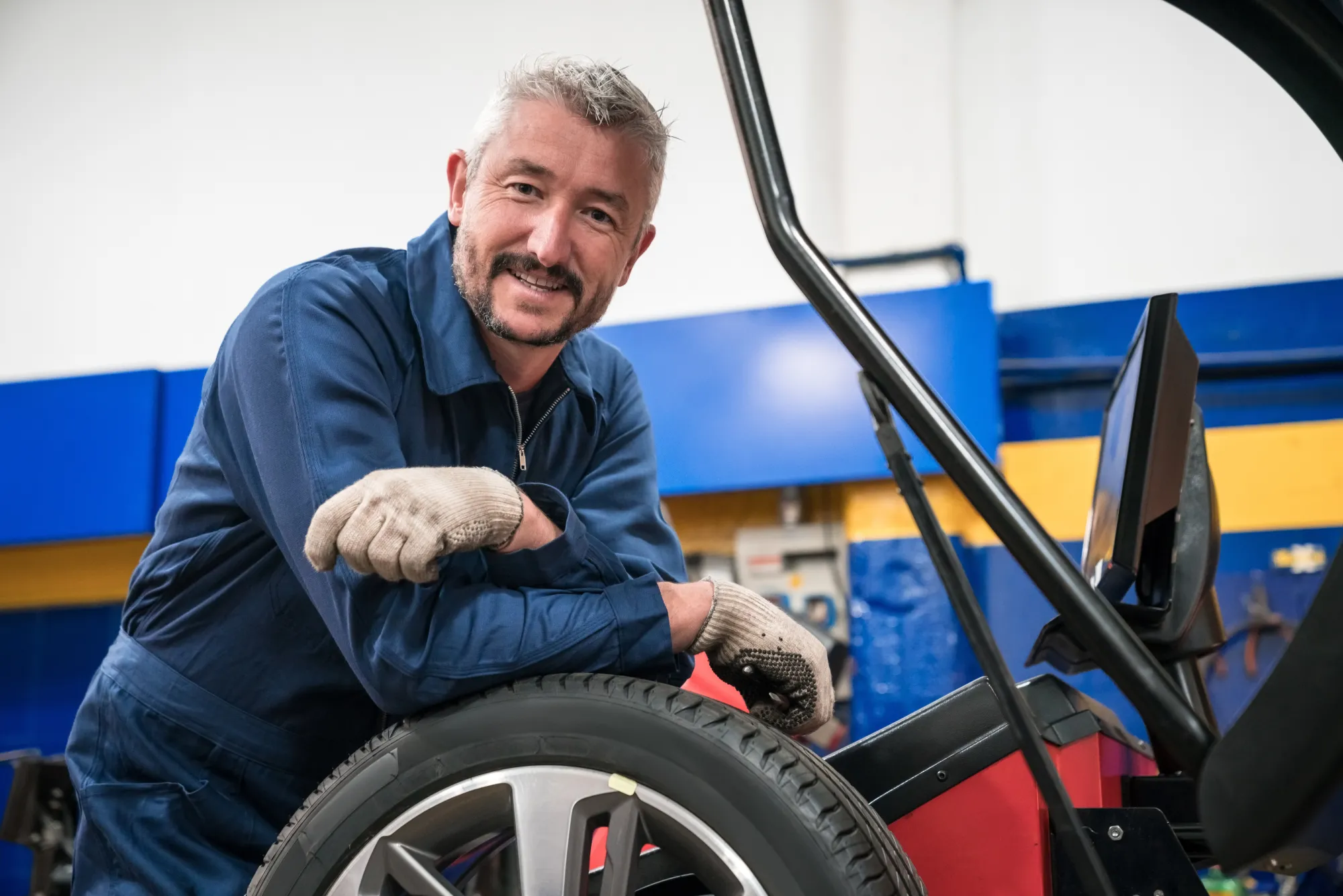 A smiling mechanic in blue coveralls and work gloves leans on a tire and alloy wheel loaded in a tire-changing machine inside an auto shop.