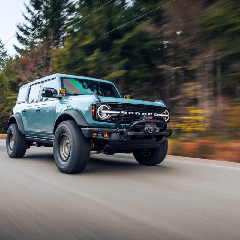 A light blue Ford Bronco SUV with large off-road tires driving along a paved road lined with blurred evergreen and autumn trees.
