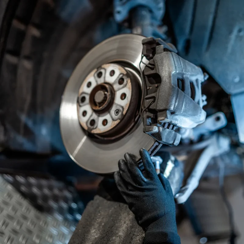 A gloved mechanic's hand works on the brake caliper and rotor of a car with its wheel removed in a repair shop.
