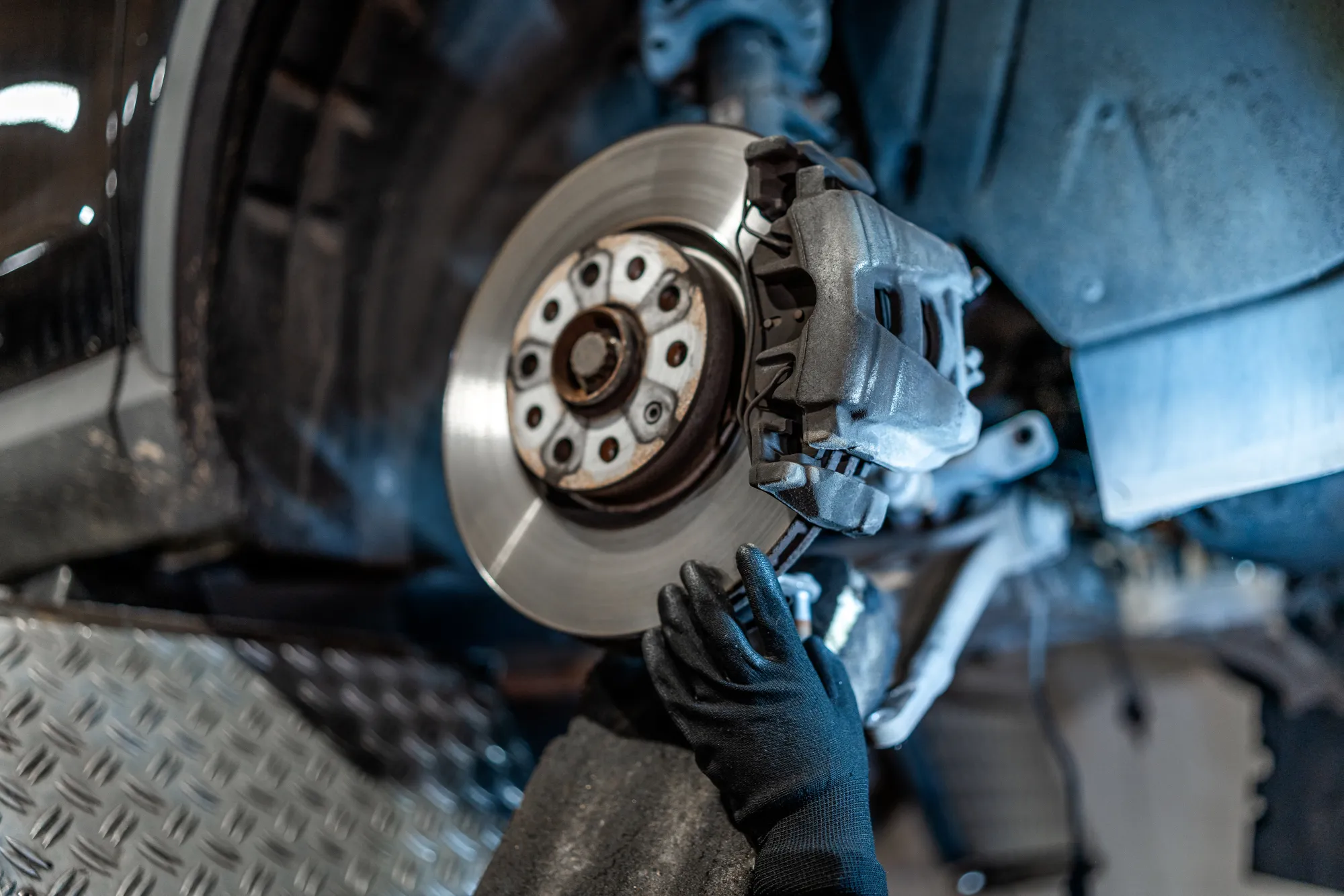A gloved mechanic's hand works on the brake caliper and rotor of a car with its wheel removed in a repair shop.